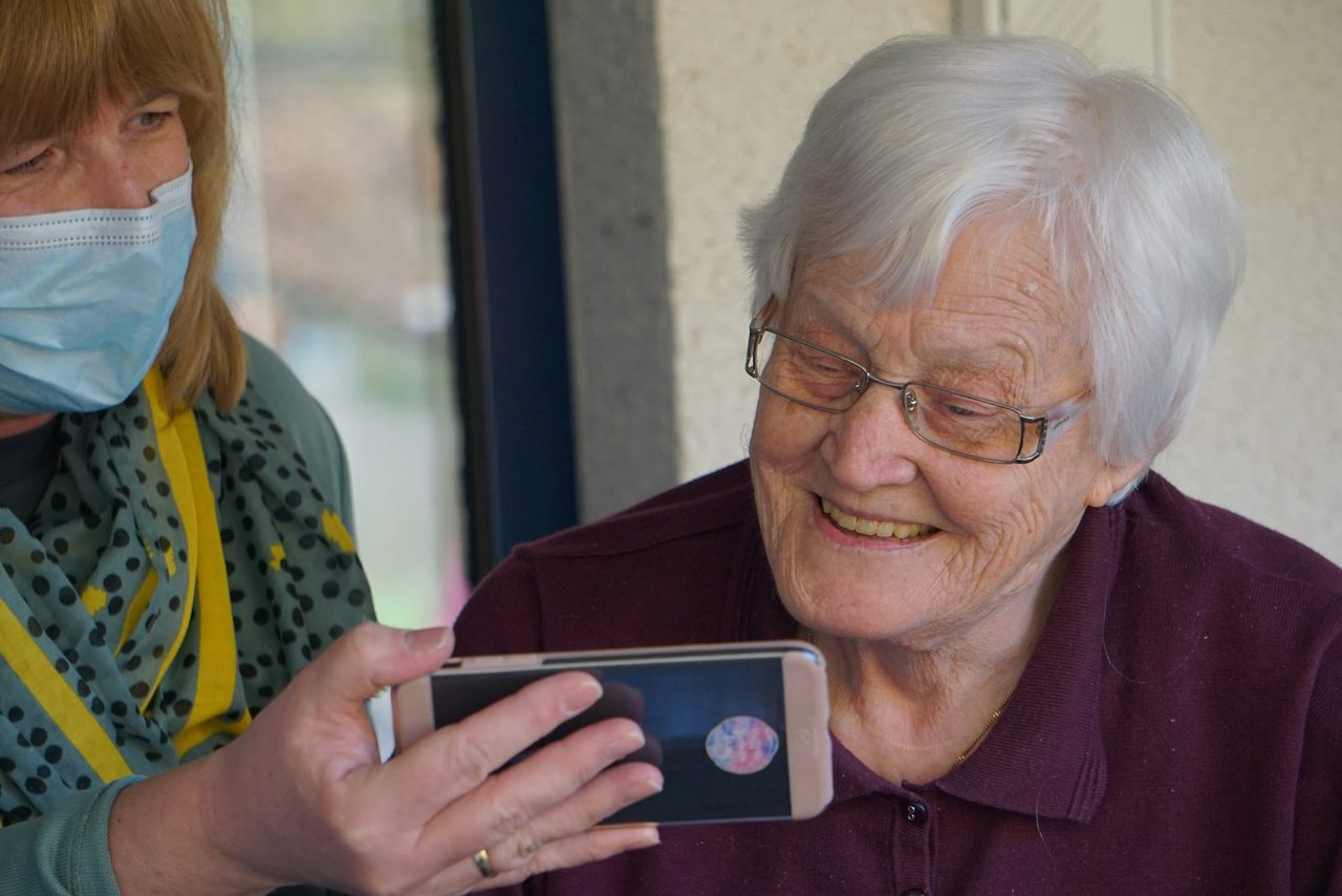 An older patient smiling as a carer helps her access her consent on a phone